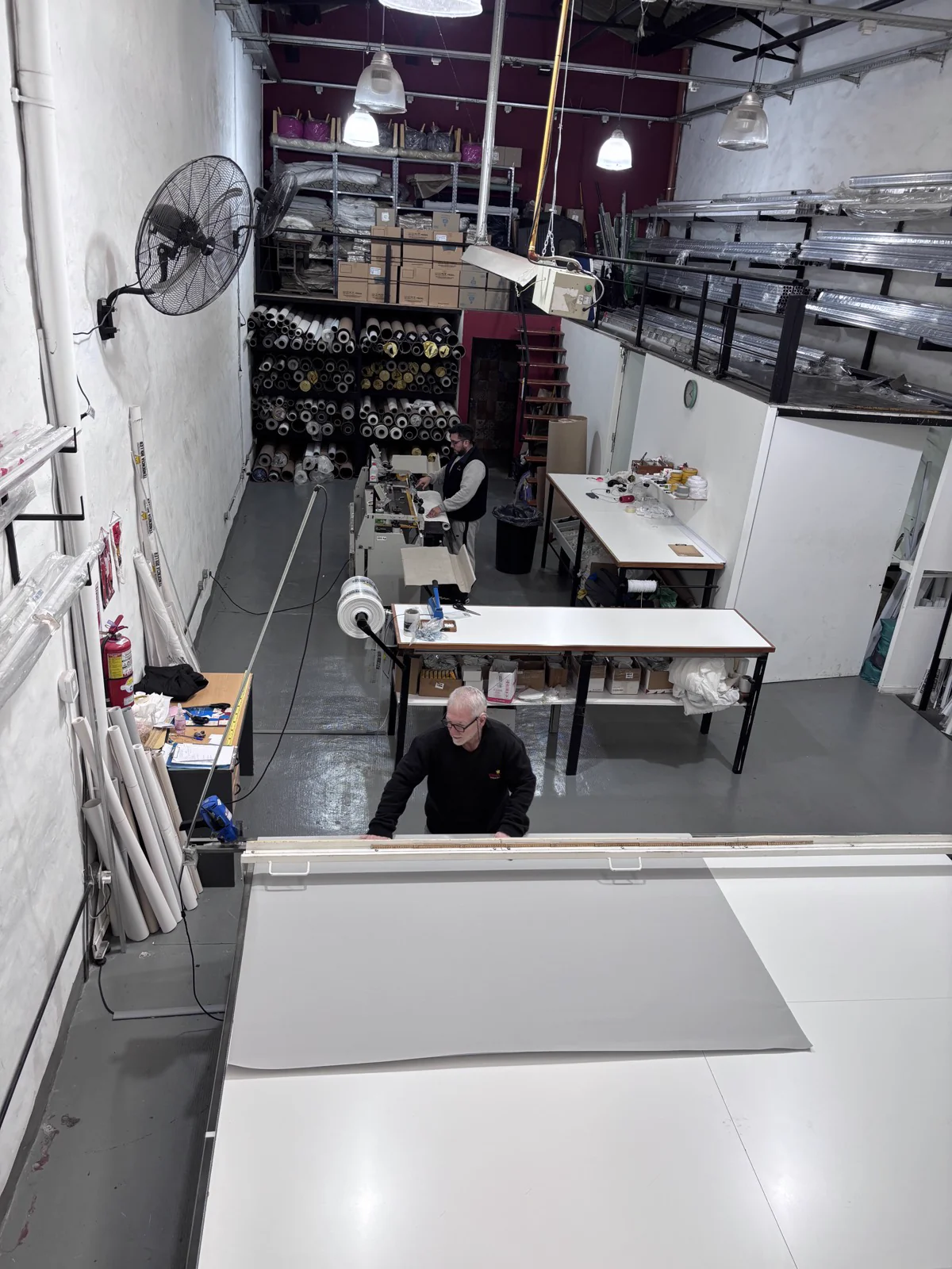 Vista del taller desde el entrepiso: rollos de tela, mesas de corte y dos operarios trabajando un paño a medida.